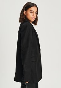 Blazer noir avec épaules structurées, fermeture à un bouton et poches latérales. Le tissu semble épais et texturé. Design ajusté, coupe droite.