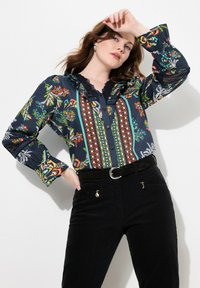 Blusa floreale blu marina con motivi vivaci, colletto rifinito in pizzo e chiusura a bottoni. Abbinata a pantaloni in corduroy neri con dettagli zip.