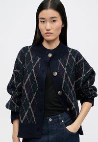 Cardigan marine avec un motif argyle en vert, rouge et beige. Dispose d'un col haut, d'une fermeture à boutons et de manches amples. Texture tricotée.