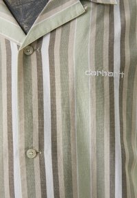 Chemise à manches courtes avec des rayures vert clair, beige et blanche. Elle présente un logo sur la poitrine et un col classique avec une fermeture à un bouton.