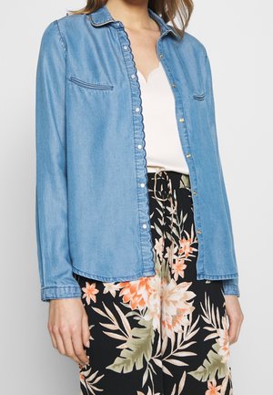 Femme portant une chemise en jean bleu clair par-dessus un top blanc et un pantalon noir avec un imprimé floral orange et vert, debout sur un fond neutre.