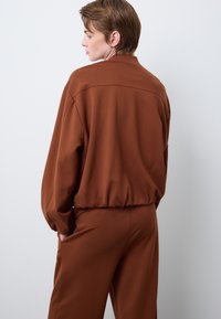 Felpa marrone, ampia, con colletto alto, orlo elasticizzato e maniche lunghe, realizzata in un tessuto morbido e strutturato.