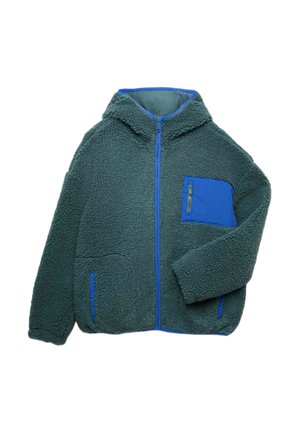 Junge steht, trägt eine grüne Fleecejacke mit blauem Taschen- und Reißverschluss, dunkelblaue Hose und weiße Sneakers mit braunen Sohlen.