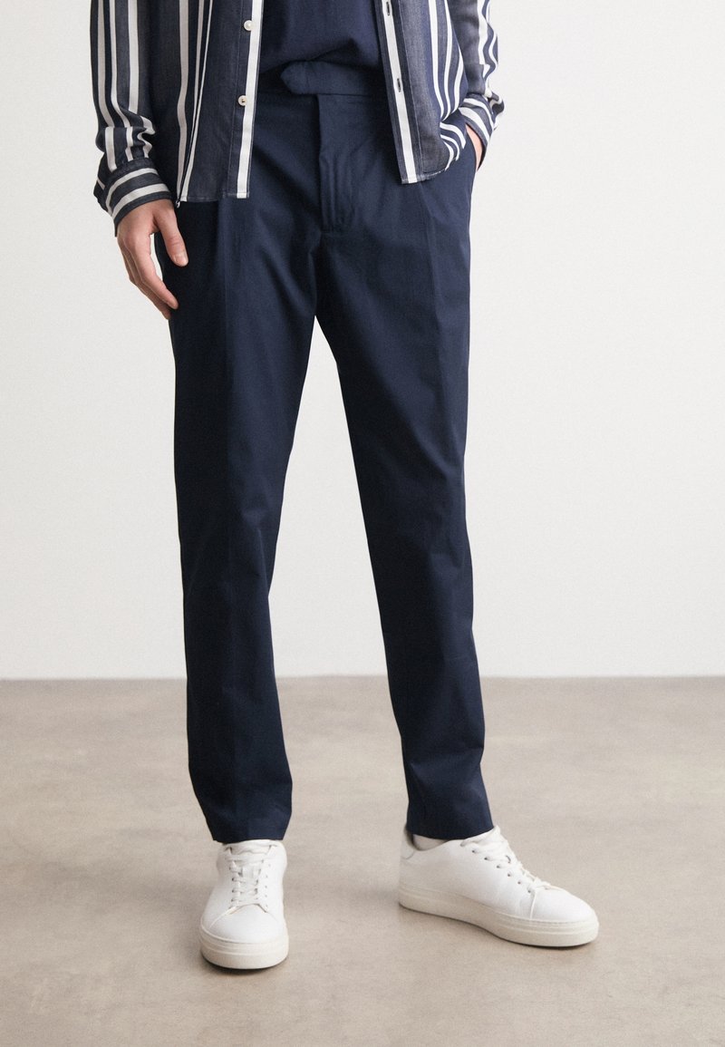 Man med marinblå byxor, vita sneakers och en randig skjorta med händerna i fickorna som står på ett slätt golv.