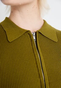 Olívazöld bordázott zip-záras felső, hajtott gallérral, fényes cipzárral és puha tapintással. Közelkép a nyakrészről.