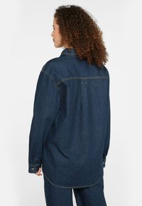 Dark denim shirt with long sleeves, a collar, and back pleat detail. Features visible stitching and a relaxed fit.