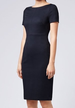 Kobieta ubrana w dopasowaną, krótkorękawową, czarną sukienkę do kolan z okrągłym dekoltem, stojąca na tle jasnego, jednolitego tła.