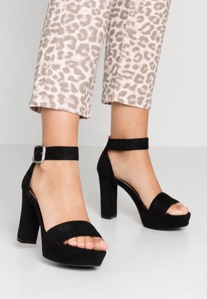 Voeten in zwarte suède plateauhakken met enkelriempjes, gecombineerd met beige cropped broeken met luipaardprint.