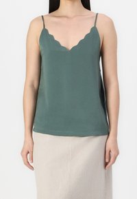 Camisola verde teal con scollatura a scallop, spalline sottili e una consistenza liscia. Abbinata a una gonna leggera beige a righe verticali.
