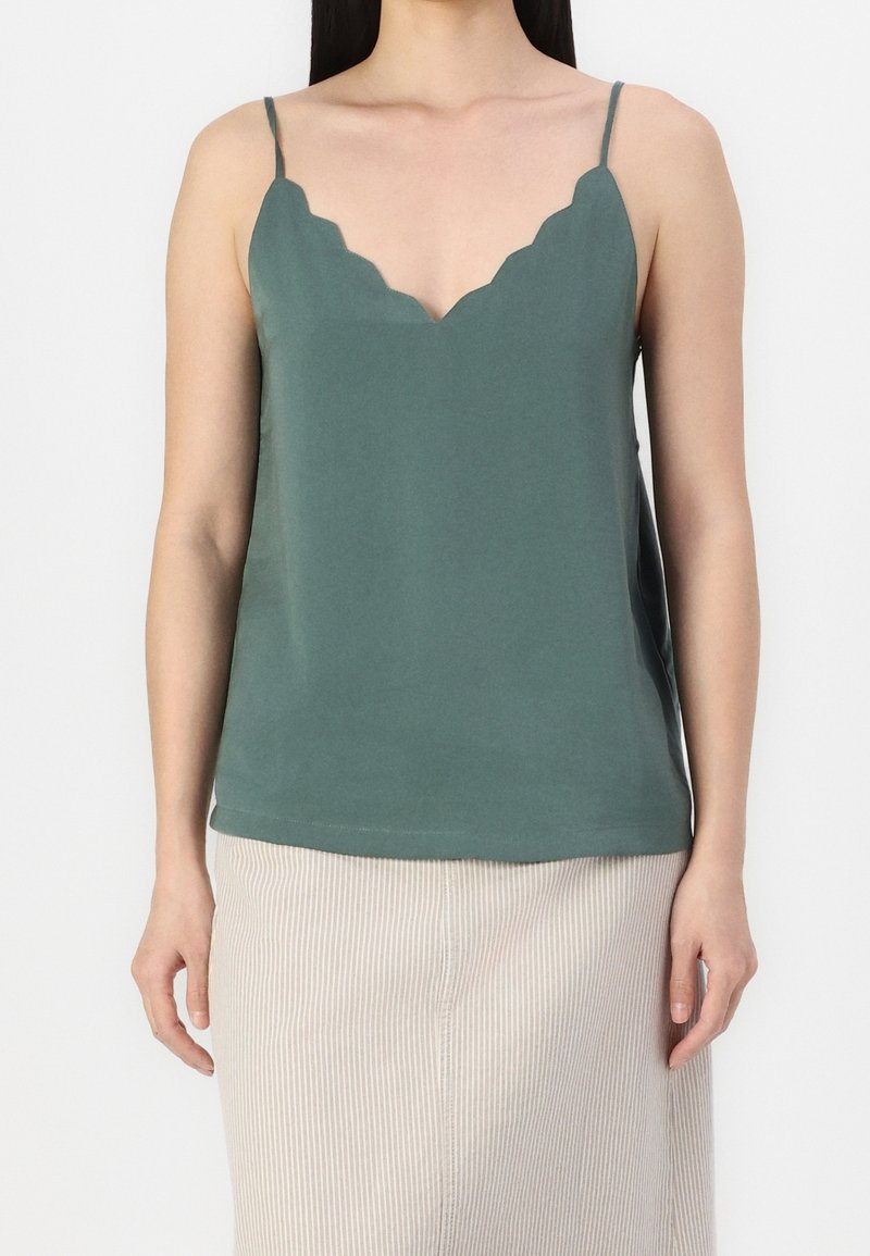 Camisola verde teal con scollatura a scallop, spalline sottili e una consistenza liscia. Abbinata a una gonna leggera beige a righe verticali.