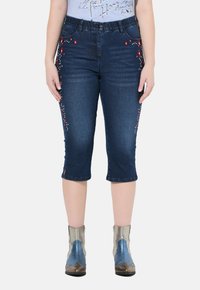 Jeansowe spodnie capri w ciemnoniebieskim kolorze, z czerwonym i białym haftem kwiatowym po bokach, z mankietem na końcach i detalami kieszeni.