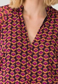 Mønstret bluse i lilla, med et geometrisk design med rosa og orange detaljer. Stof har en let, struktureret kvalitet. V-udskæring.