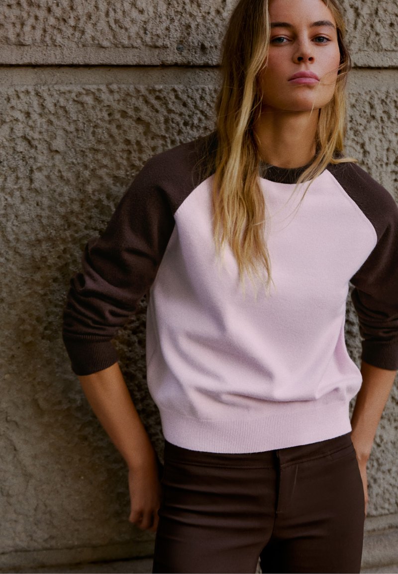 Jeune femme aux longs cheveux blonds se tenant contre un mur en pierre texturé, vêtue d’un pull raglan rose et marron et d’un pantalon foncé.