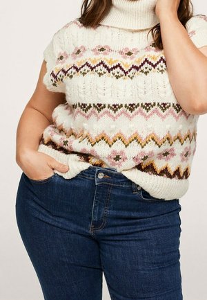 Mujer que lleva un suéter blanco de cuello alto sin mangas de punto con patrones de zigzag y flores de colores metido dentro de unos vaqueros azul oscuro.