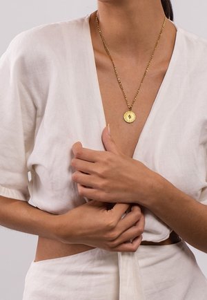 Femme portant un haut cache-cœur et une jupe en lin blanc, tenant le tissu près de la taille, avec un collier pendentif en or.