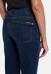 Jeans in denim blu scuro con una texture liscia, dotati di tasche posteriori, cuciture visibili e un'etichetta con il brand sopra la tasca destra.