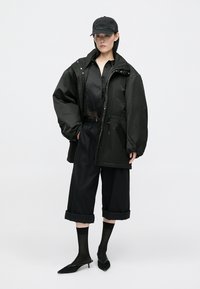 Preveliki crni parka s sjajnom završnom obradom, složen preko prozirne crne košulje, u kombinaciji s skraćenim crnim hlačama i špicastim crnim petama.