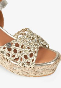 Sandale plateforme métallique dorée tissée avec une semelle en jute tressé, présentant un large design de brides entrecroisées et une semelle intérieure lisse couleur tan.