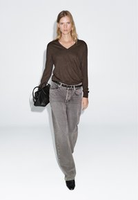 Brown v-neck sweater, gray straight-leg jeans, black belt, and black handbag. Model wears pointed black heels, standing against a white background.
