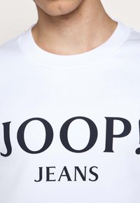 Sweat-shirt blanc en coton avec une grande impression noire indiquant "JOOP! JEANS" en police audacieuse et moderne. Le col est en tricot côtelé.