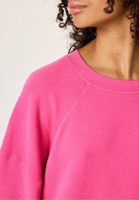 Roza dukserica s okruglim izrezom i raglan rukavima, koja ima mekanu teksturu i minimalne detalje šivanja.