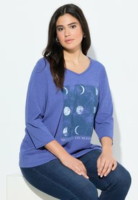 Jonge vrouw met lang donker haar, gekleed in een paarse top met driekwartmouwen met een print van maanfasen en donkerblauwe jeans, zittend op een witte blok.