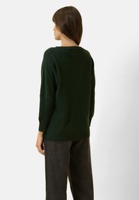 Vrouw die met de rug naar de camera staat, gekleed in een donkergroene trui met lange mouwen en donkergrijze broek, tegen een effen lichte achtergrond.