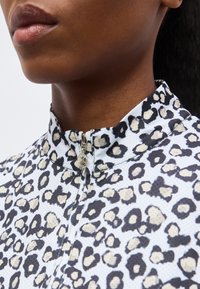 Κοντινό πλάνο ενός ατόμου που φοράει μπλούζα με μπεζ και μαύρο λεοπάρ σχέδιο, με ψηλό γιακά και λεπτομέρειες φερμουάρ.