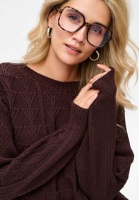 Bordowy sweter dzianinowy z teksturowanym trójkątnym wzorem, z ribbami na rękawach i szerokim dekoldem. Duże okulary w kolorze żółwi dopełniają wygląd.