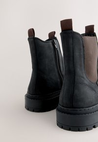 Paar schwarze Stiefeletten mit dicken Sohlen, seitlichen Reißverschlüssen und braunen elastischen Einsätzen sowie Zuglaschen vor hellem Hintergrund.