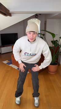 Gorro de punto beige, sudadera crema con cremallera hasta el pecho y gráfico "CLUB", pantalones negros y zapatillas plateadas. Ambiente interior con plantas.