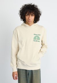 Hanorac crem cu buzunar frontal tip kangaroo, având un imprimeu grafic verde "Ellesse 1959 Club di Corsa", croială lejeră și manșete ribbate.