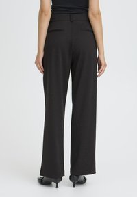 Donna che indossa pantaloni neri a vita alta e a gamba larga con un sottile motivo a pois e scarpe con tacco kitten neri, in piedi su uno sfondo bianco.