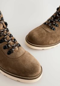 Botas de tobillo de ante marrón con costuras blancas en contraste, ojetes negros y cordones con patrón. Punta redondeada y diseño de suela resistente.