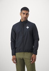 Svart lett jakke med høy krage, glidelås og elastiske mansjetter. Har en hvit logo på venstre brystområde.