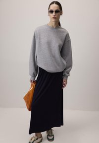 Felpa grigia oversize, gonna maxi blu navy, borsa a tracolla arancione e sneakers beige-verdi. Tessuti morbidi, vestibilità rilassata e design a strati.