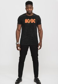 Black short-sleeve t-shirt with distressed texture features bold red and yellow "AC/DC" logo. Paired with black slim-fit pants and shiny black shoes.