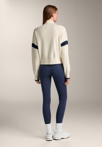 Cremfarbener Cropped-Pullover mit marineblauen Streifen, weicher Textur. Kombiniert mit marineblauen Leggings und weißen Sneakern, die gerippte Bündchen und Socken haben.