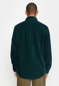 Donkergroen corduroy shirt met lange mouwen, knoopmanchetten en een rugnaaddetail. Beschikt over een verticale ribbels structuur en een afgeronde zoom.