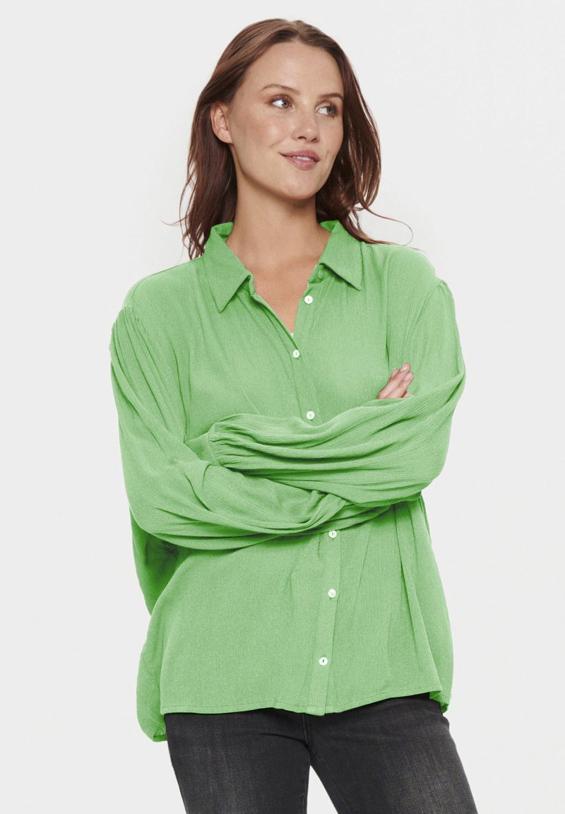 Camicia verde con bottoni, realizzata in un tessuto strutturato, con colletto, vestibilità ampia e maniche lunghe. I jeans neri completano l'outfit.