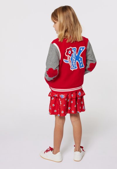 Veste varsity rouge avec des manches grises, ornée d'un patch logo Kenzo bleu et rouge. Associée à une robe rouge à motif floral avec volants et des baskets blanches.