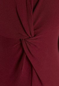 Haut bordeaux avec texture côtelée, présentant un détail torsadé à la taille. Tissu lisse au design ajusté, mettant en valeur la silhouette.