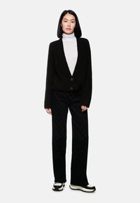 Schwarze, flauschige Strickjacke mit tiefem V-Ausschnitt und vorderen Knöpfen, kombiniert mit schwarz gemusterten, weit geschnittenen Hosen und klobigen Sneakers mit weißer Sohle.