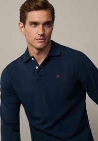 Marineblå langermet polo skjorte laget av bomull, med klassisk krage og tre knapper foran, samt en rød brodert logo på brystet.