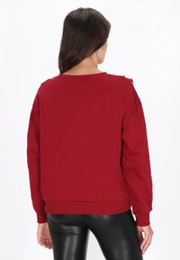 Piros pulóver kerek nyakkivágással, hosszú ujjakkal, bordás mandzsettákkal és enyhén bő fazonnal. Vállán díszítő részletekkel.