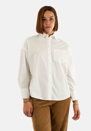 Camisa blanca de botón hecha de algodón, con un bolsillo en el pecho, que presenta mangas largas y un corte relajado, combinada con pantalones de pana marrones.