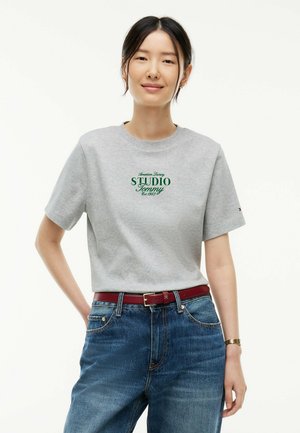 Donna che indossa una maglietta grigio chiaro con testo verde che dice ""STUDIO Tommy"" e jeans blu con cintura rossa su sfondo bianco.