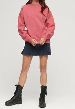 Frau trägt einen rosa langärmeligen Pullover, einen marineblauen plissierten Rock und schwarze Schnür-Kampfstiefel und steht vor einem einfarbigen Hintergrund.