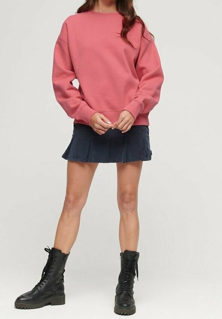 Femme portant un sweat-shirt rose à manches longues, une jupe plissée bleu marine et des bottes de combat noires à lacets, debout devant un fond uni.