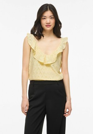 Mladá žena s čiernymi vlasmi, ktorá nosí bledožlté tielko bez rukávov s volánmi a čierne nohavice s vysokým pásom, stojaca pred bielym pozadím.